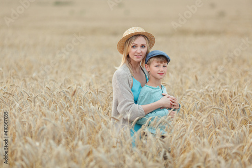 Mom with a straw hat and blue dress hugs her son in a cap standing in a wheat field. Summer holidays in the village © Diana