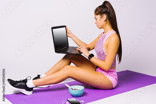 A young sports girl with a laptop eats muesli and watches a workout video while sitting on the mat. White background.