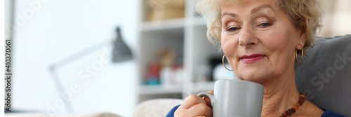 Portrait of lovely beautiful elderly female holding cup of tasty tea and reading interesting book. Smiling attractive woman relaxing on couch in living room. Leisure and self education concept photo