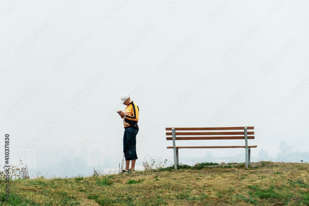 A man stands on a hilltop with a phone in his hands against a background of thick fog. An empty bench and a young man standing next to it. The concept of loneliness and longing.