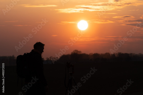 The silhouette of a person against the background of the sun and the beautiful sky. The outline of a man standing next to a tripod and a camera on the background of the sunset.