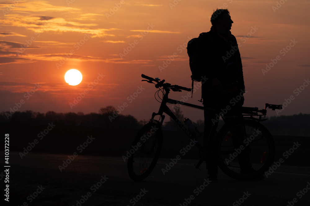 Fototapeta premium The silhouette of a cyclist against the background of the sun and the beautiful sky. The outline of a man standing next to his bicycle against the background of the sunset.