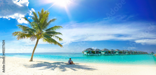 Beautiful beach with white sand, turquoise ocean, palm and blue sky with clouds on sunny day. Man sits on sand in shade of palm tree. Sun is at its zenith. Summer tropical landscape, panoramic view.