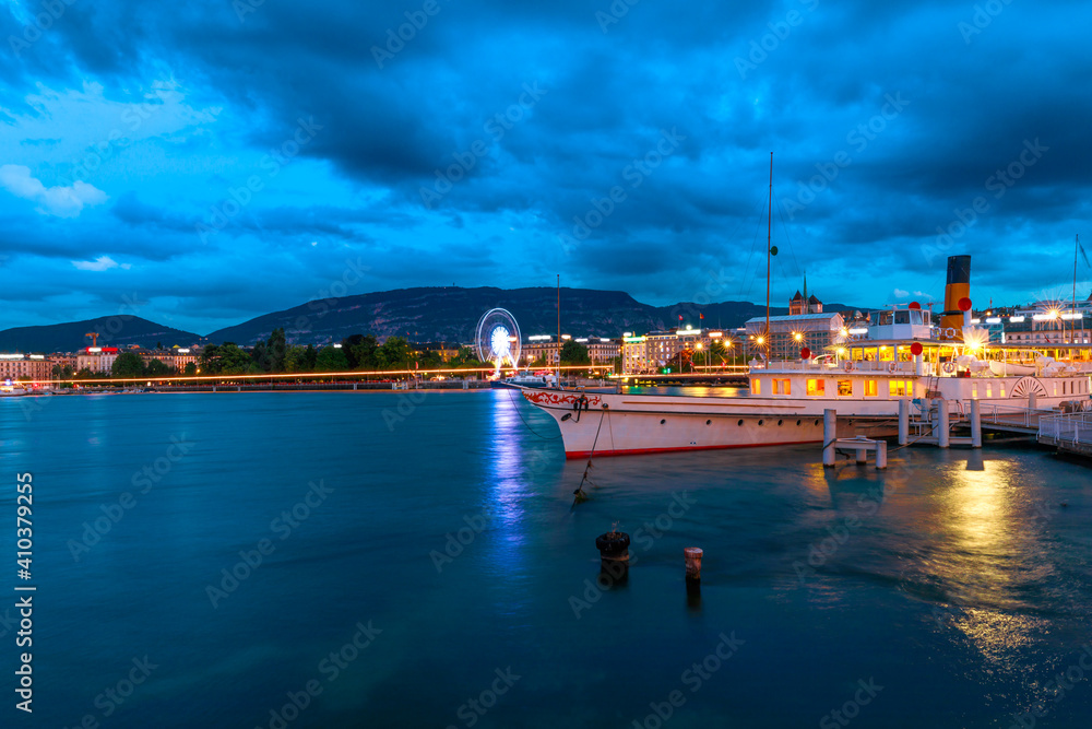Naklejka premium Dramatic stormy sky of Geneva urban cityscape and Swiss Alps reflecting in Lake Leman, Switzerland in French Swiss. Pier in Quai du Mont-Blanc ferry terminal at dusk, illuminated in the evening.