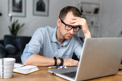 Thoughtful young man in modern glasses and smart casual wear is looking on laptop screen and solving some tasks. A guy works online from home