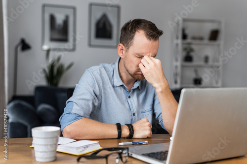 Overworked young entrepreneur in tired from work with a laptop, holds on to the bridge of the nose his eyes closed. Stressed guy works remotely at home feels eyes strain