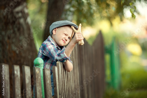 little boy a bully in a cap and a plaid shirt sitting on a wooden fence shooting a slingshot
