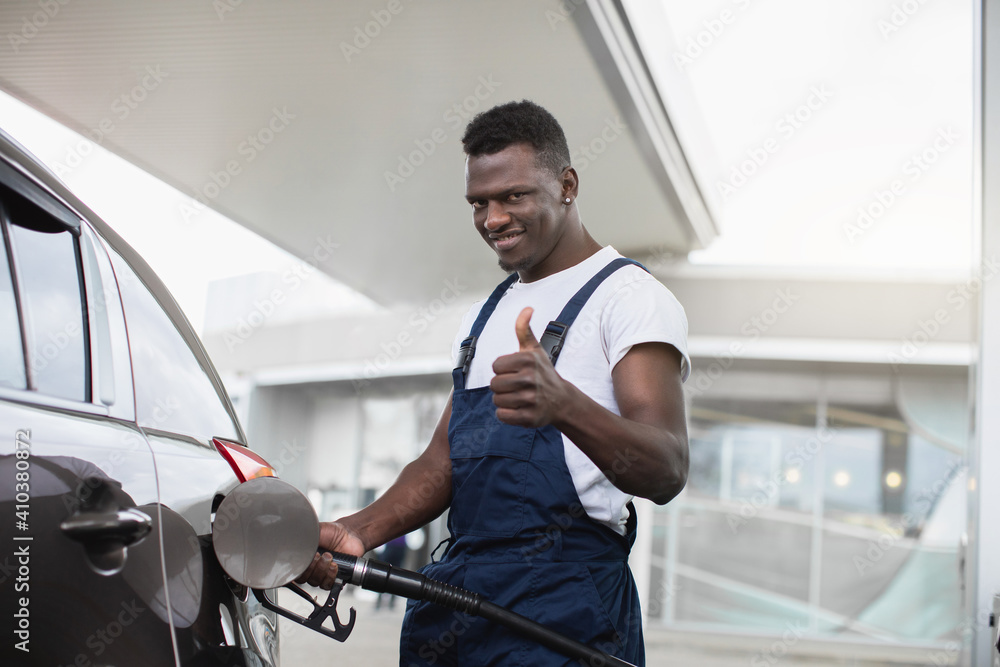 Smiling African American guy, worker at the gas station, dressed on white t-shirt and blue overalls, showing his thumb up, while filling a luxury car Smiling African American guy, worker at the gas station, dressed on white t-shirt and blue overalls, showing his thumb up, while filling a luxury car