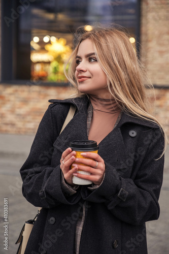 Blonde woman in dark coat drinks coffee in to go cup. Dark cold autumn evening. Europeaan city lights on the background. Space for text photo