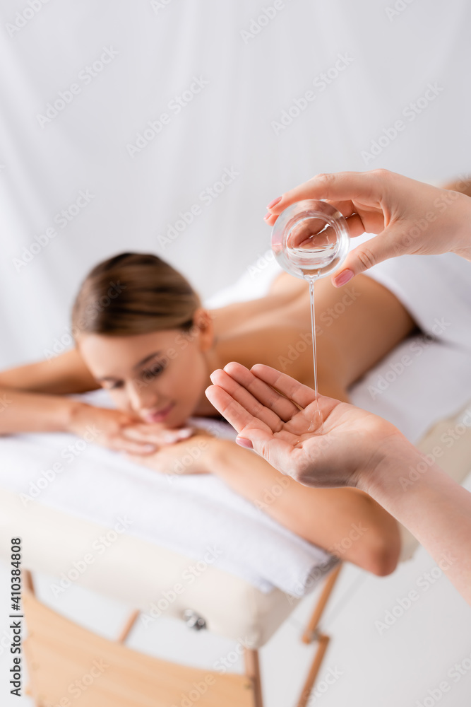 © LIGHTFIELD STUDIOS - masseur holding glass container and pouring oil on hand near woman resting on massage table and blurred background