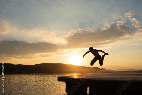 A child jumps from a pier into a lake during sunset. Summer rest. Active spending time. Vacation and Holidays. Focus on the sunset.
