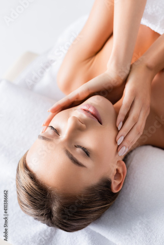 pleased young woman with closed eyes touching face and lying on massage table in spa salon