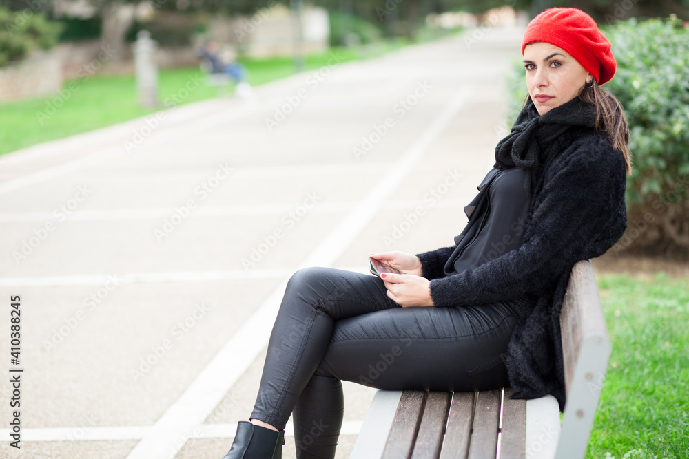 Fototapeta premium Ragazza mora vestita di nero con cuffietta rossa è seduta in u panchina in un parco