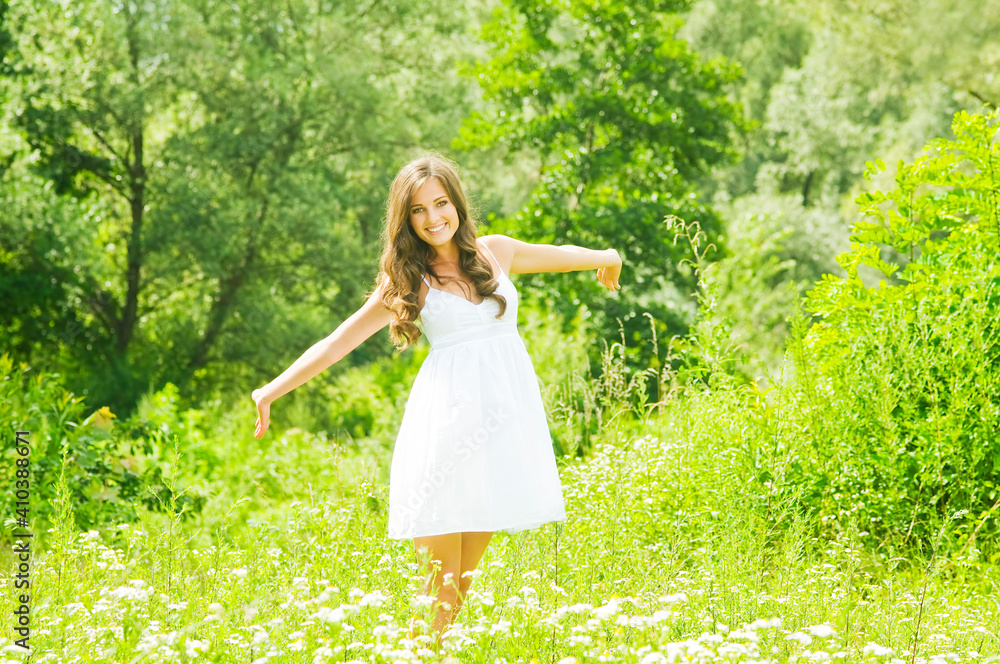 Smiling young woman in white dress against background of summer green park. Beautiful healthy happy girl enjoying freedom outside in nature.