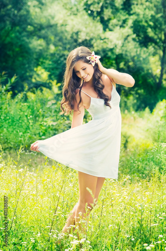 Smiling young woman in white dress with flower against background of summer green park. Beautiful healthy happy girl enjoying freedom outside in nature.