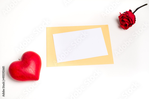Top view on a white and blank gift card on a sand-colored envelope, with a red love heart on its left and a red rose on the right, on a white background. Valentine's day and love 