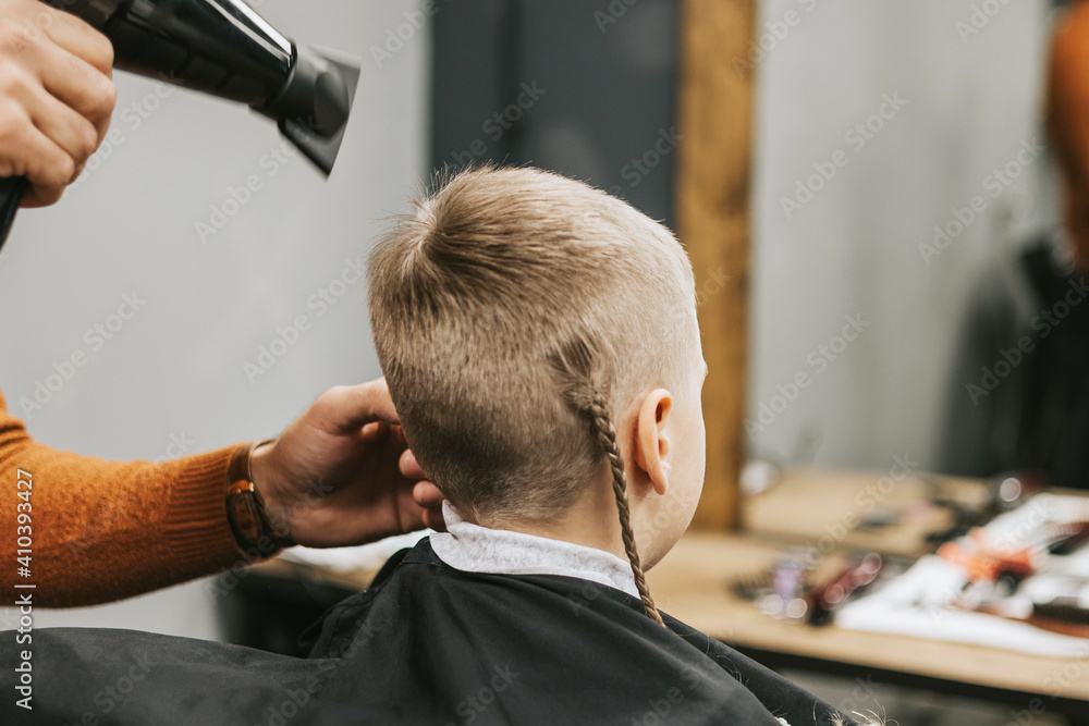 Fototapeta premium the process of hair styling a blond boy with with hair dryer and comb in a chair in a barbershop salon, a barbershop concept for men and boys