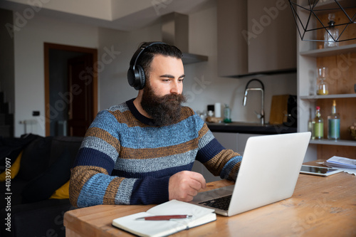 Young man at home at table working on computer with headphones - Concept of smartworking in lockdown - Millennials in a business meeting with his colleagues remotely - Male attends an online course photo