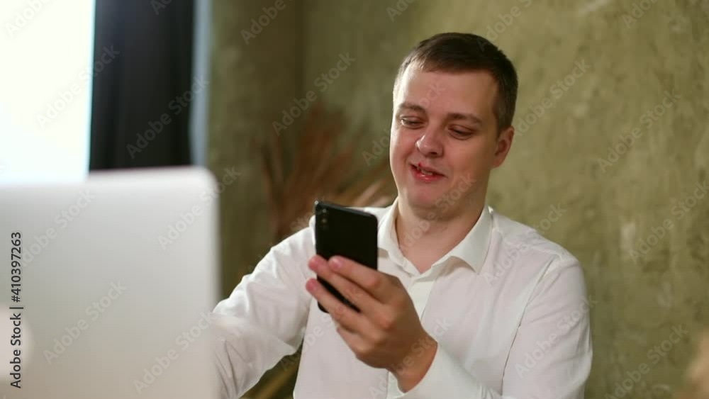 Young smiling business man in elegant white shirt smiling using smartphone looking at screen typing message. Goods news. Internet communication app. Freelance lifestyle. Home office. Online shopping. Young smiling business man in elegant white shirt smiling using smartphone looking at screen typing message. Goods news. Internet communication app. Freelance lifestyle. Home office. Online shopping.