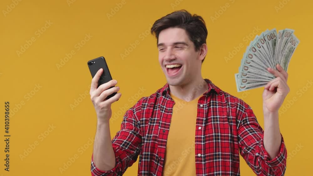 Overjoyed happy young man 20s in casual red shirt posing isolated on yellow background in studio. People lifestyle concept. Using mobile cell phone hold fan of cash money in dollar banknotes screaming Overjoyed happy young man 20s in casual red shirt posing isolated on yellow background in studio. People lifestyle concept. Using mobile cell phone hold fan of cash money in dollar banknotes screaming