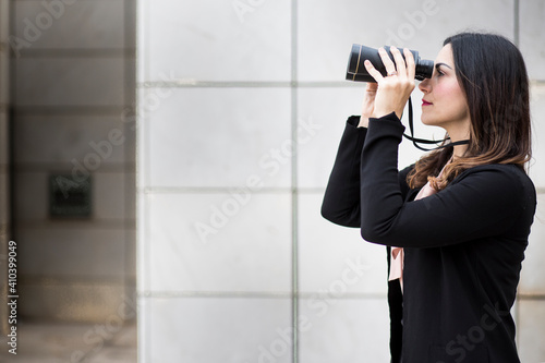 Manager donna mora,  vestita in tailleur nero, utilizza dei binocoli per guardare lontano, isolata su sfondo muro bianco di mattonelle rettangolari photo