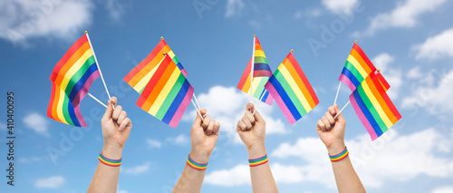 lgbt  same-sex relationships and homosexual concept - close up of male hands wearing gay pride awareness wristband holding rainbow flags over blue sky and clouds background