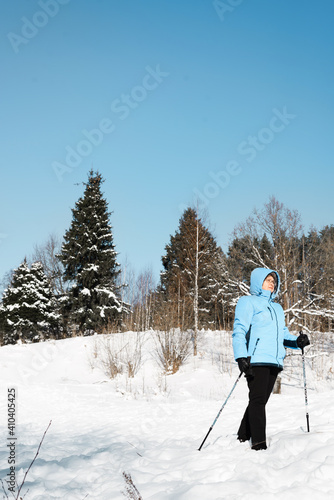 Senior woman in glasses with Nordic walking poles on the background of the winter forest. Active lifestyle retirement concept. Sunny winter day.