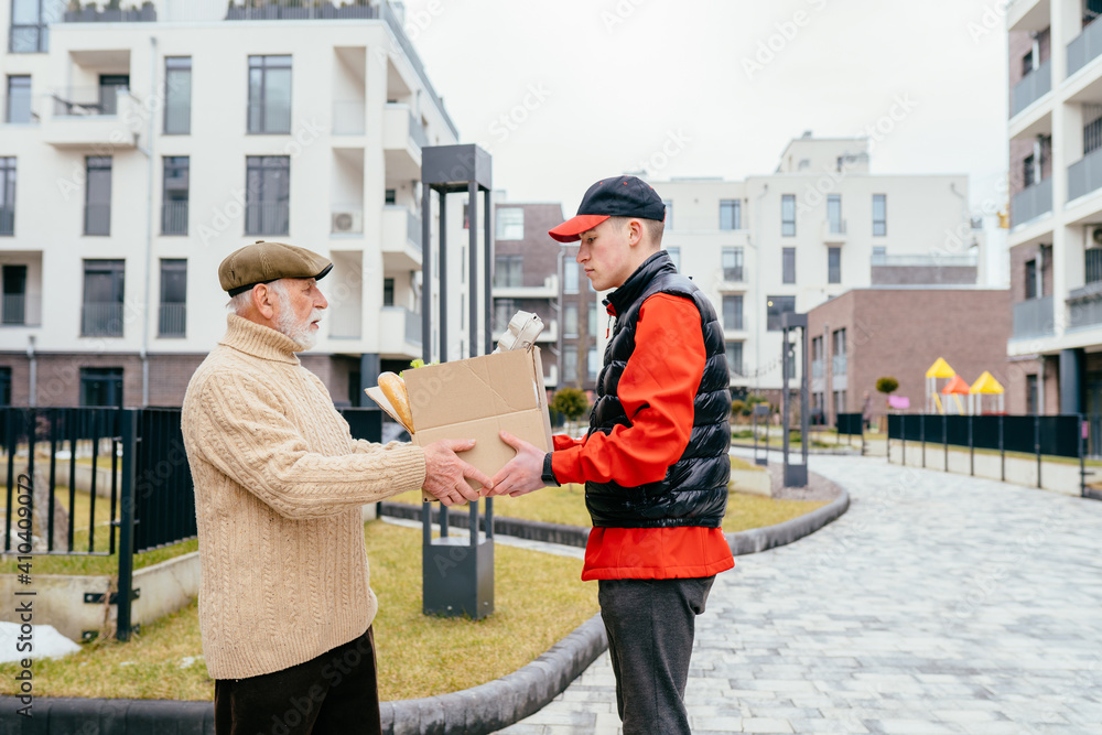 Profile shot of an elderly senior man taking to a food delivery male with a grocery box outdoor with building on background. Meal basket as social help and support.