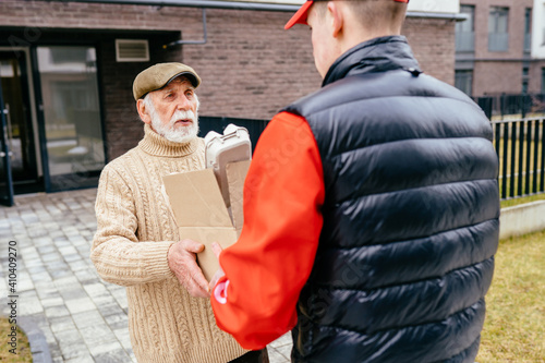Young male courier in uniform delivering parcel box with meal to senior man outdoor with doorway of apartment house on background. Fresh groceries and food delivery for elderly people.