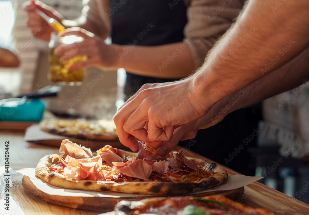 Close-up of a man and a woman's hands are preparing pizza in the kitchen in a restaurant, laying out the meat and oiling. on a sunny day