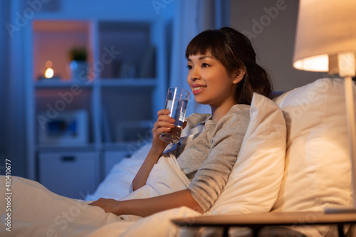 people, bedtime and thirst concept - happy smiling asian woman drinking water at night in bed at home