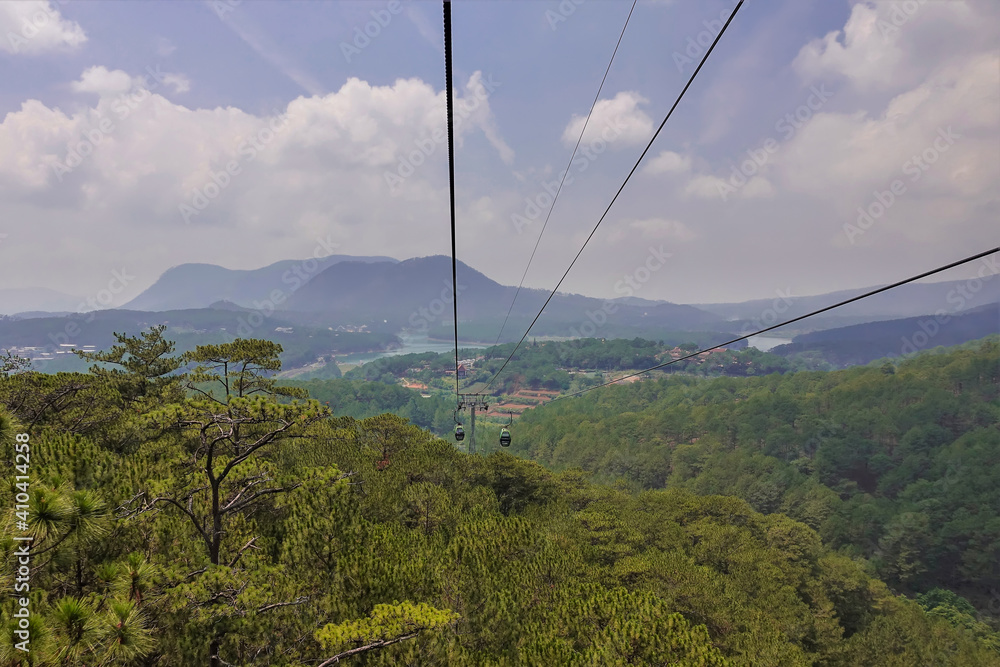 The cable car cabins move over the coniferous forest. Silhouettes of mountains and a lake are visible in the distance. Clouds in the blue sky. Vietnam. Dalat The cable car cabins move over the coniferous forest. Silhouettes of mountains and a lake are visible in the distance. Clouds in the blue sky. Vietnam. Dalat