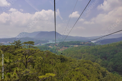 The cable car cabins move over the coniferous forest. Silhouettes of mountains and a lake are visible in the distance. Clouds in the blue sky. Vietnam. Dalat