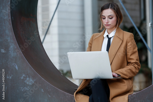 фотографія manager donna bionda con i capelli a caschetto, camicia bianca , cappotto beige e  cravatta nera, lavora al computer in esterni con espressione seria