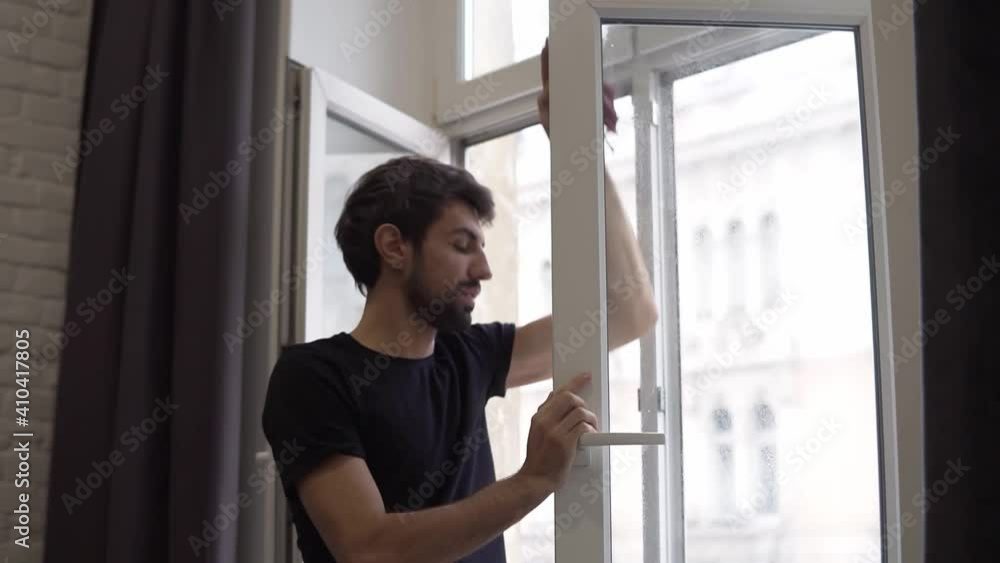 Man is cleaning window, cleans surface and uses a rag, standing in modern apartment Man is cleaning window, cleans surface and uses a rag, standing in modern apartment