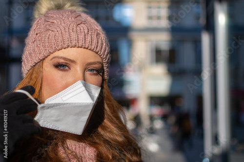 Woman with blue eyes, ginger hair, and a woollen hat during pandemic isolation as part of the prevention of the COVID-19 diseases spread at city street. Removing protection face mask (FFP-2) from face © Lukas