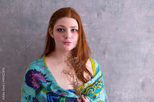 Medium horizontal frontal view of beautiful young woman with long bright red hair wearing a blue-green wrap and holding a dry flower © Anne Richard