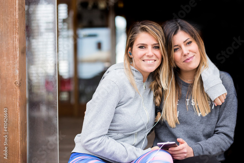 amiche in abbigliamento casual si abbracciano felici sedute vicine a un chiosco e insieme ascoltano la musica dello smartphone dalle cuffie auricolari photo