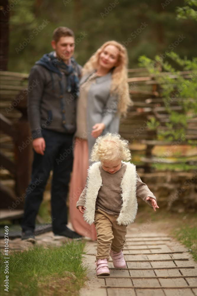 Fototapeta premium A curly little girl with blond hair in a warm waistcoat and beige trousers walks with her parents in the courtyard of a house in the village. Concept of a happy family. Village