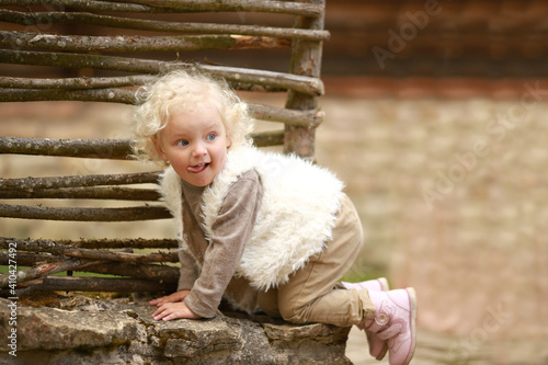 Cheerful girl with curly white hair in a vest crawling next to the fence of her house.