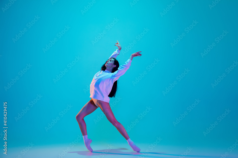 Inspired. Young and graceful ballet dancer isolated on blue studio background in neon light. Art, motion, action, flexibility, inspiration concept. Flexible caucasian ballet dancer, moves in glow.