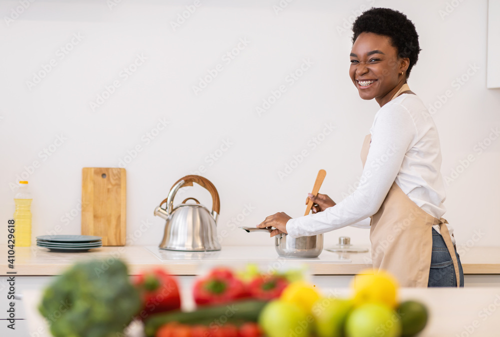 Fototapeta premium Happy African Woman Cooking Dinner In Kitchen, Smiling To Camera