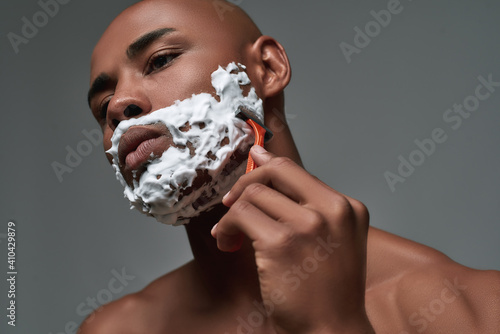 Close up portrait of handsome young african american man with foam applied on his face using steel razor for shaving isolated over gray background
