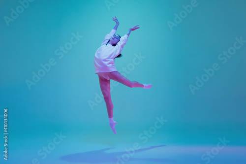 Flying high. Young and graceful ballet dancer isolated on blue studio background in neon light. Art, motion, action, flexibility, inspiration concept. Flexible caucasian ballet dancer, moves in glow.