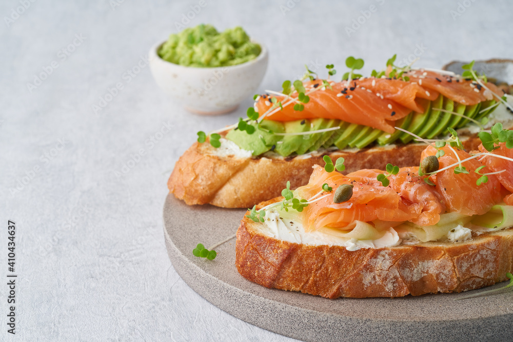 Two open sandwich, toast with salmon, cream cheese, avocado, cucumber slices on white concrete table. Morning healthy breakfast with fish and cup of drink, side view, copy space Two open sandwich, toast with salmon, cream cheese, avocado, cucumber slices on white concrete table. Morning healthy breakfast with fish and cup of drink, side view, copy space