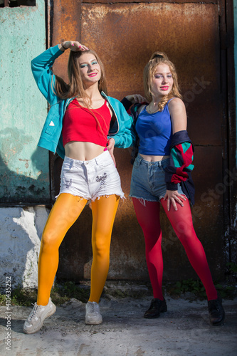 Two funny girls in bright clothes in the style of the nineties are standing against the background of a rusty door. Russian village girls. © Светлана Лазаренко