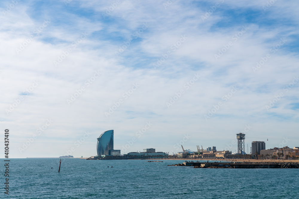 BARCELONA, SPAIN, FEBRUARY 3, 2021: The famous W hotel, known as hotel vela. Sunny winter day. View from inside the water. In the background we can see the cargo area of the port and the cable car. BARCELONA, SPAIN, FEBRUARY 3, 2021: The famous W hotel, known as hotel vela. Sunny winter day. View from inside the water. In the background we can see the cargo area of the port and the cable car.
