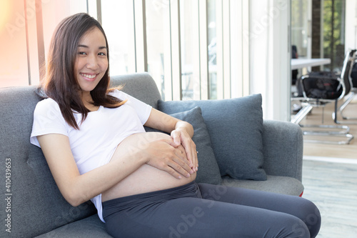 Elegant Relaxed Asian Young Happy Pregnant Woman keeping hand on belly and sitting on couch in living room.