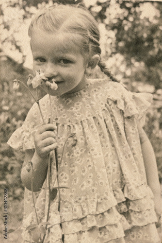 Latvia - CIRCA 1930s: Portrait of girl playing with flower in garden. Vintage coloured Art Deco era photography