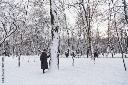 Aged smiling woman standing near to a big tree in a snow-covered winter park. Snowy winter